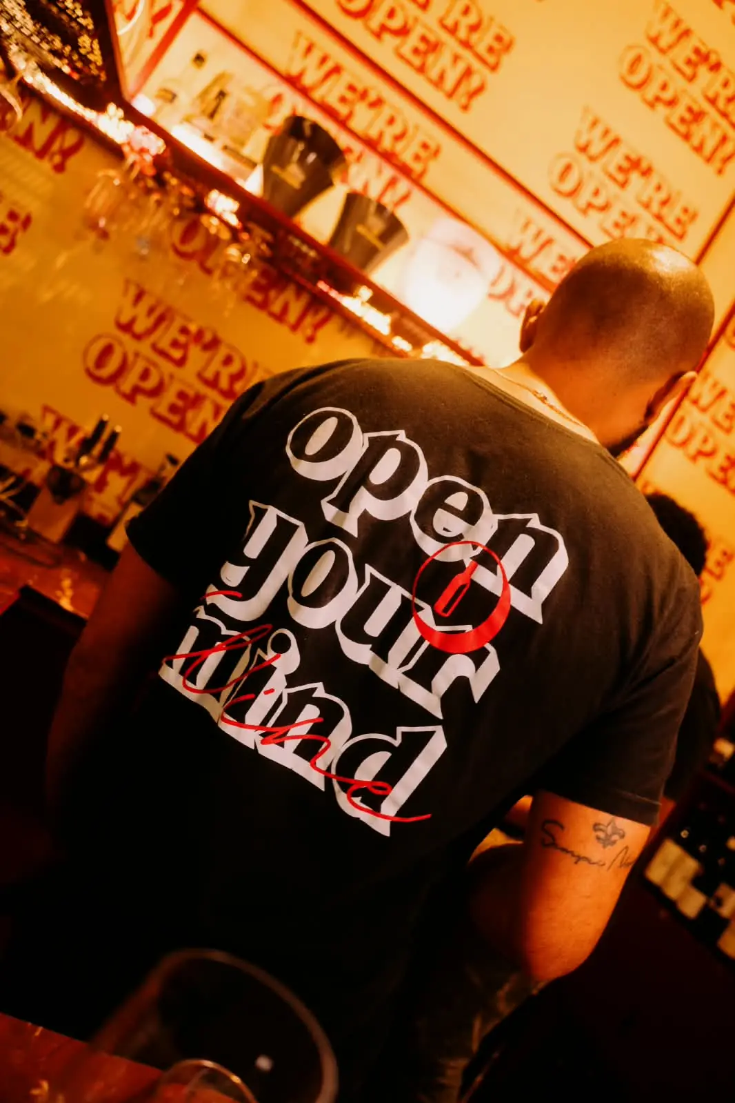 Atendente de costas com camiseta "Open your mind" diante da parede "We're open!"