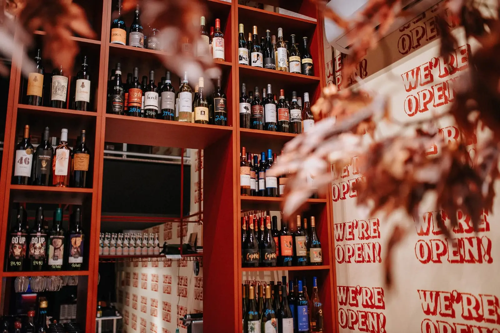 Prateleiras vermelhas cheias de garrafas e a parede "We're open!" ao lado de uma árvore